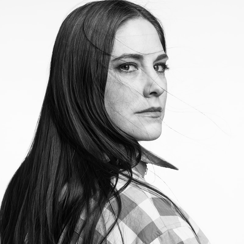 A black and white portrait of Jessica, a white woman with long, dark, straight hair.  A few flyaways blow in front of her face, which emotes a serious expression.  She sits in profile looking over her right shoulder and directly into the camera lens.  She wears a baggy, checkered man's shirt with a popped collar.